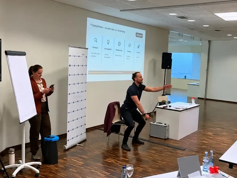 Tim Hermle in der Hocke vor einer Präsentationsleinwand in einem Seminarraum, links eine Teilnehmerin am Flipchart