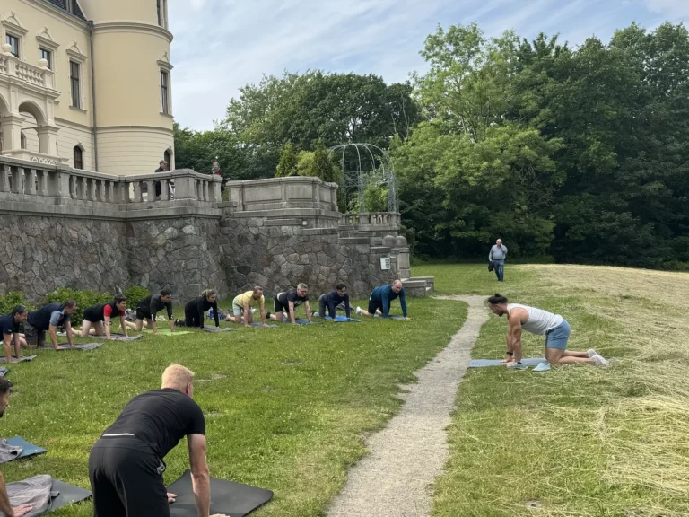 Eine Firmen-Yogagruppe übt im Vierfüßlerstand auf Matten entlang der Schlossmauer im Park von Schloss Ralswiek